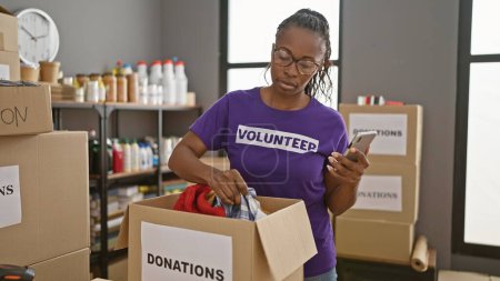 Photo for African american woman volunteering, sorting donations in a warehouse, while checking her phone. - Royalty Free Image