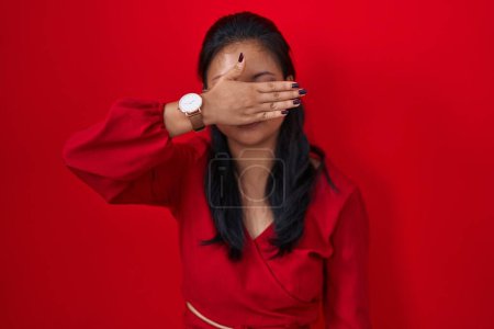 Téléchargez les photos : Asiatique jeune femme debout sur fond rouge couvrant les yeux avec la main, l'air grave et triste. concept de non-visibilité, de dissimulation et de rejet - en image libre de droit