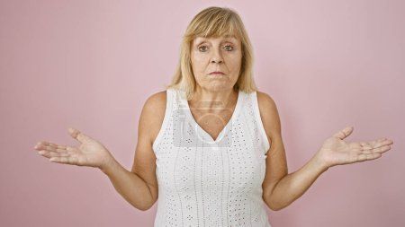 Clueless middle age blonde woman, expression filled with doubt and confusion, standing indifferent over isolated pink background, lost in thought, arms relaxed