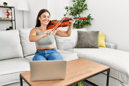Foto de Joven hermosa mujer hispana que tiene clase de violín en línea sentado en el sofá en casa - Imagen libre de derechos
