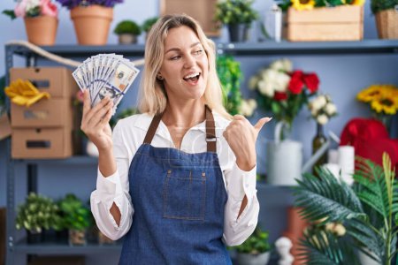Photo for Young blonde woman working at florist shop holding dollars pointing thumb up to the side smiling happy with open mouth - Royalty Free Image
