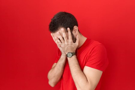 Foto de Young hispanic man wearing casual red t shirt with sad expression covering face with hands while crying. depression concept. - Imagen libre de derechos