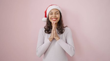 Téléchargez les photos : Jeune belle femme hispanique priant souriant confiant portant un chapeau de Noël sur fond rose isolé - en image libre de droit