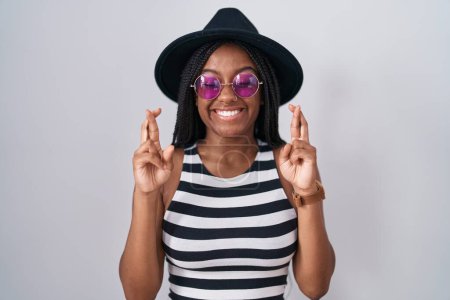 Téléchargez les photos : Jeune afro-américain avec des tresses portant un chapeau et des lunettes de soleil geste doigt croisé souriant avec espoir et les yeux fermés. chance et concept superstitieux. - en image libre de droit