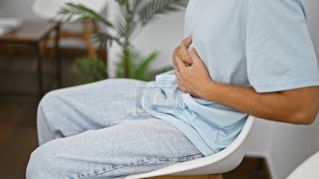 Foto de Hombre hispano con dolor de estómago sentado en una moderna sala de espera interior. - Imagen libre de derechos