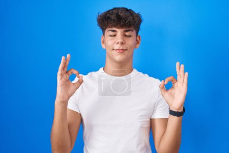 Téléchargez les photos : Adolescent hispanique debout sur fond bleu détendu et souriant avec les yeux fermés faisant geste de méditation avec les doigts. concept de yoga. - en image libre de droit