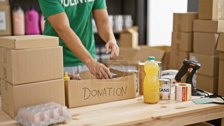Photo for A young man prepares a donation box with food items in a warehouse setting, reflecting charity and community service. - Royalty Free Image