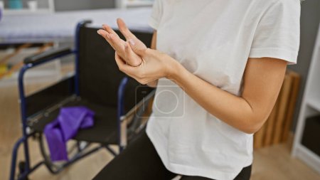 Téléchargez les photos : Une jeune femme effectue des exercices de main dans une salle de clinique de réadaptation avec un fauteuil roulant visible. - en image libre de droit