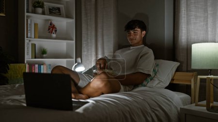Photo for Enthralled young hispanic teenager, comfortably sitting on his bed, engrossed in watching a movie on his laptop, radiating a positive vibe in his cozy bedroom at night, making every pixel count. - Royalty Free Image