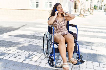 Téléchargez les photos : Jeune belle femme hispanique parlant sur smartphone assis sur fauteuil roulant à la rue - en image libre de droit