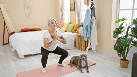 Foto de Mujer de pelo gris de mediana edad entrena a su perro mientras hace ejercicio sus piernas en el dormitorio, una opción de estilo de vida para mantenerse en forma y saludable - Imagen libre de derechos