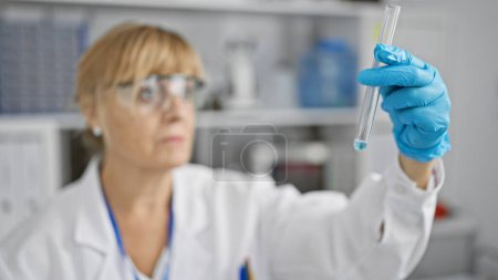 Photo for Dedicated middle age blonde woman scientist, safely clad in glasses and gloves, meticulously measures test tube contents in a bustling lab. - Royalty Free Image