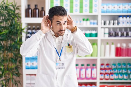 Téléchargez les photos : Un bel homme hispanique qui travaille à la pharmacie en essayant d'entendre les deux mains sur le geste de l'oreille, curieux de commérages. problème auditif, sourd - en image libre de droit