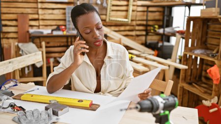 Photo for Attractive african american woman carpenter in serious conversation on smartphone, reading important document amidst her carpentry workshop - Royalty Free Image
