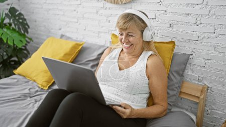 Téléchargez les photos : Bénie femme blonde d'âge moyen se détendre dans sa chambre confortable, assis sur le lit avec ordinateur portable, profiter de la musique à travers des écouteurs, rayonnant de bonheur et de confiance - en image libre de droit
