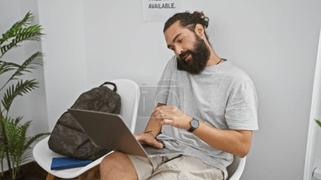 Téléchargez les photos : Jeune homme hispanique barbu utilisant un ordinateur portable dans une salle intérieure moderne, illustrant un environnement de travail décontracté. - en image libre de droit