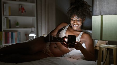 Foto de Mujer afroamericana usando lencería usando smartphone acostado en la cama en el dormitorio - Imagen libre de derechos