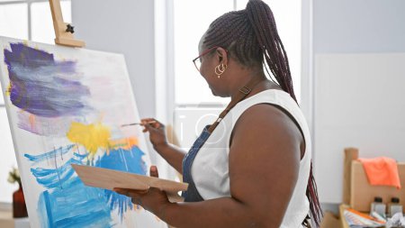 Téléchargez les photos : Artiste afro-américaine focalisée, avec des tresses et des lunettes, dessinant sérieusement sur toile au studio d'art d'intérieur - en image libre de droit