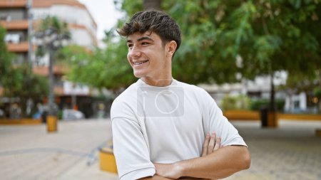 Photo for Cheerful young hispanic teenager confidently standing in city park, arms crossed, happily looking to the side, enjoying nature's smile. - Royalty Free Image
