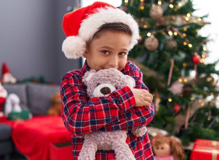 Foto de Adorable niño hispano abrazando oso de peluche de pie junto al árbol de Navidad en casa - Imagen libre de derechos