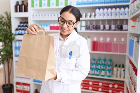 Téléchargez les photos : Jeune pharmacienne arabe souriante confiante tenant son sac à provisions à la pharmacie - en image libre de droit