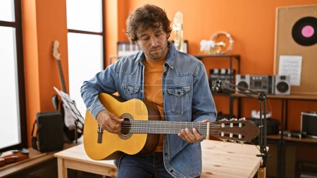 Foto de Hombre hispano guapo tocando la guitarra en un estudio de música con paredes naranjas y equipo moderno. - Imagen libre de derechos