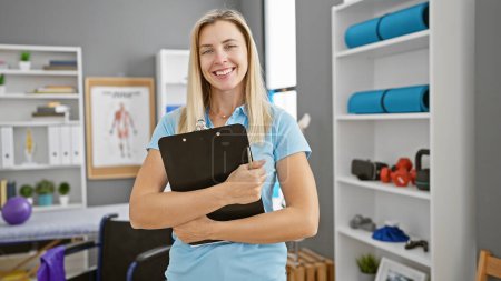 Photo for A smiling young blonde woman clinician stands confidently in a rehab clinic with exercise equipment. - Royalty Free Image