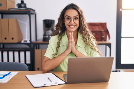 Photo for Young hispanic woman working at the office wearing glasses praying with hands together asking for forgiveness smiling confident. - Royalty Free Image