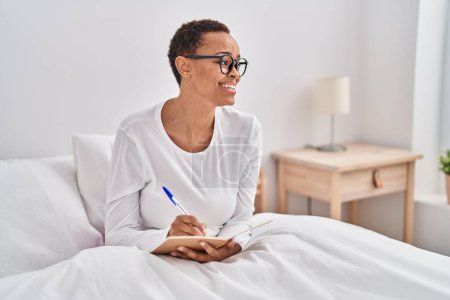 Téléchargez les photos : Femme afro-américaine écrivant sur un cahier assis sur le lit dans la chambre - en image libre de droit