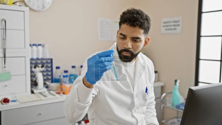 Foto de Un latino enfocado en una bata de laboratorio examina un tubo de ensayo en un ambiente de laboratorio moderno. - Imagen libre de derechos