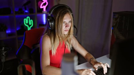 Photo for Focused young caucasian woman gaming on computer in a dark room wearing headphones. - Royalty Free Image