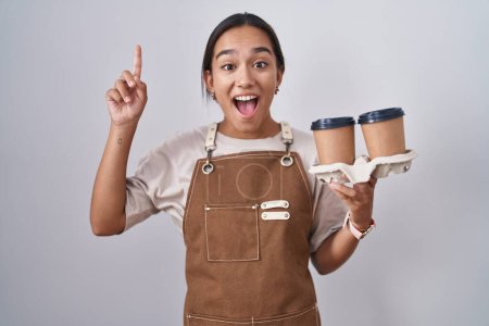Photo for Young hispanic woman wearing professional waitress apron holding coffee smiling amazed and surprised and pointing up with fingers and raised arms. - Royalty Free Image