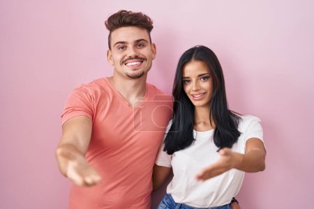 Photo for Young hispanic couple standing over pink background smiling friendly offering handshake as greeting and welcoming. successful business. - Royalty Free Image