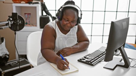Téléchargez les photos : Femme afro-américaine souriante, musicienne confiante, composant une chanson pleine d'âme dans un studio de musique, absorbée dans son monde musical. - en image libre de droit
