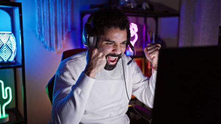 Téléchargez les photos : Homme hispanique excité avec une barbe dans une salle de jeux sombre célébrant une victoire la nuit. - en image libre de droit