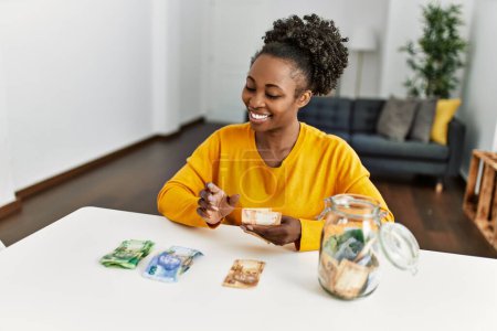 Foto de Mujer afroamericana sentada en la mesa separando billetes de rand en casa - Imagen libre de derechos