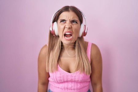 Téléchargez les photos : Jeune femme blonde écoutant de la musique à l'aide d'écouteurs en colère et fous hurlant frustrés et furieux, criant de colère. rage et concept agressif. - en image libre de droit