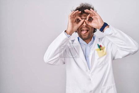 Téléchargez les photos : Jeune homme hispanique portant l'uniforme de médecin faisant un geste correct comme des jumelles sortant la langue, les yeux regardant à travers les doigts. expression folle. - en image libre de droit