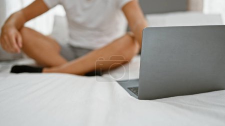 Téléchargez les photos : Un jeune homme décontracté assis sur un lit, avec un ordinateur portable ouvert devant lui dans une chambre lumineuse - en image libre de droit