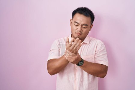 Téléchargez les photos : Jeune homme chinois debout sur fond rose souffrant de douleur aux mains et aux doigts, inflammation de l'arthrite - en image libre de droit
