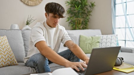 Photo for Concentrated young hispanic male student, engrossed in online education, sitting on cozy living room sofa, focused on laptop screen, relaxing at home, surrounded by technology - Royalty Free Image
