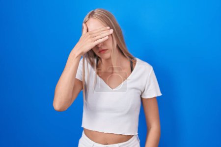 Photo for Young caucasian woman standing over blue background covering eyes with hand, looking serious and sad. sightless, hiding and rejection concept - Royalty Free Image