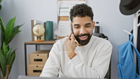 Foto de Un joven hispano sonriente con barba hablando en un teléfono móvil en un entorno de oficina moderno - Imagen libre de derechos