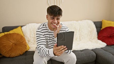 Photo for Young, handsome hispanic man smiling confidently, comfortably sitting on living room sofa, relaxing at home while enjoying using touchpad gadget, embodying a joyful expression and positive lifestyle. - Royalty Free Image