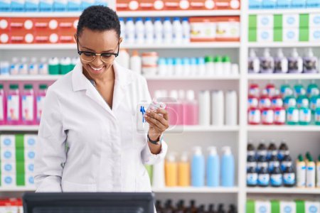 Téléchargez les photos : Afro-américaine femme pharmacien souriant confiant tenant comprimés comprimés à la pharmacie - en image libre de droit