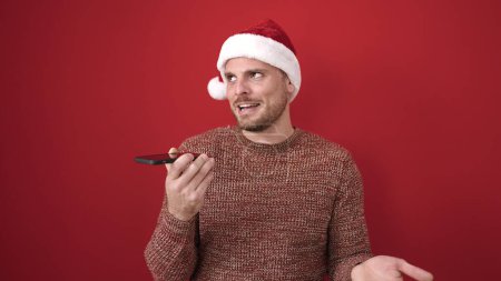 Téléchargez les photos : Jeune homme caucasien envoyant un message vocal portant un chapeau de Noël sur fond rouge isolé - en image libre de droit