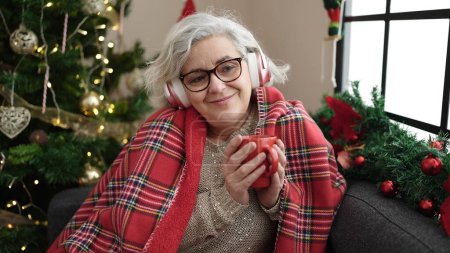 Téléchargez les photos : Femme d'âge moyen aux cheveux gris écoutant de la musique boire du café assis près de l'arbre de Noël à la maison - en image libre de droit