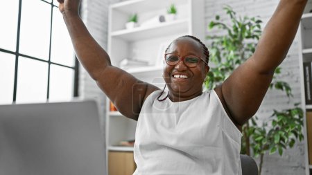 Photo for Confident african american woman, a successful business worker, raising arms in a celebrating gesture at her office desk, all smiles while working online on her laptop. - Royalty Free Image