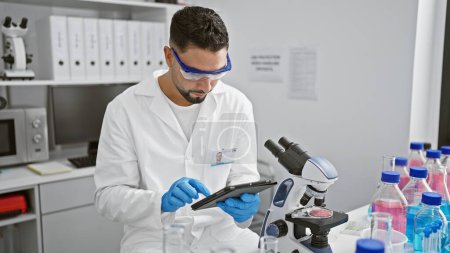 Téléchargez les photos : Homme barbu en blouse de laboratoire utilisant une tablette dans un laboratoire moderne avec microscope et échantillons. - en image libre de droit