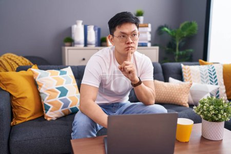 Photo for Young asian man using laptop at home sitting on the sofa thinking concentrated about doubt with finger on chin and looking up wondering - Royalty Free Image